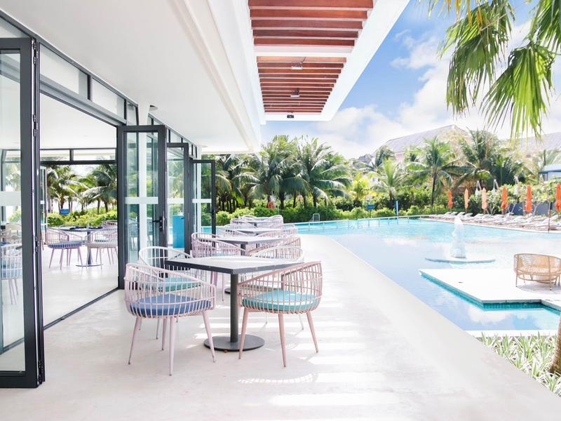 Área exterior moderna con mesas, sillas y piscina rodeada de palmeras y cielo azul.