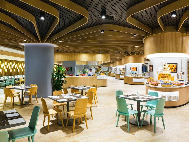 Comedor moderno y luminoso con techo de madera y varias áreas de asientos en un hotel.