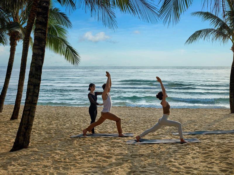 Dos personas practican yoga en la playa bajo palmeras con vista al mar tranquilo.
