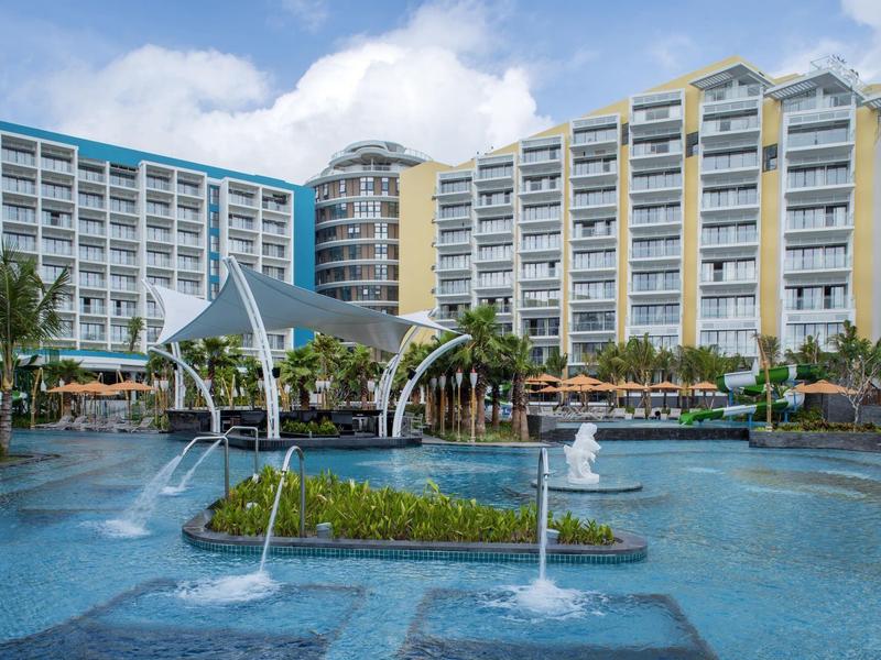 Gran piscina al aire libre con fuentes frente a un moderno edificio hotelero bajo cielo azul.