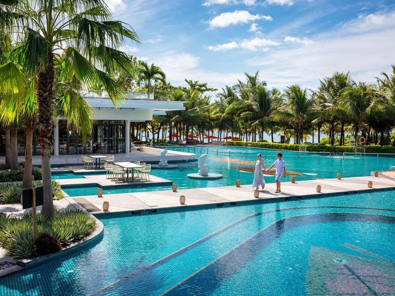 Gran piscina con áreas de agua, palmeras y tumbonas alrededor bajo un cielo azul.