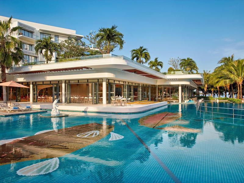 Piscina de resort moderna con bar, palmeras y agua azul clara bajo un cielo soleado.