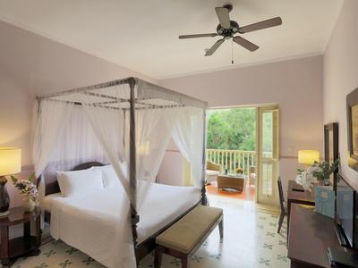 Bright hotel room with canopy bed, ceiling fan, and balcony overlooking greenery.