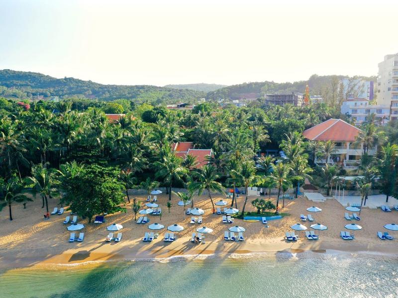 Beach with lounge chairs and palm trees in front of a hotel and wooded hills in the background.