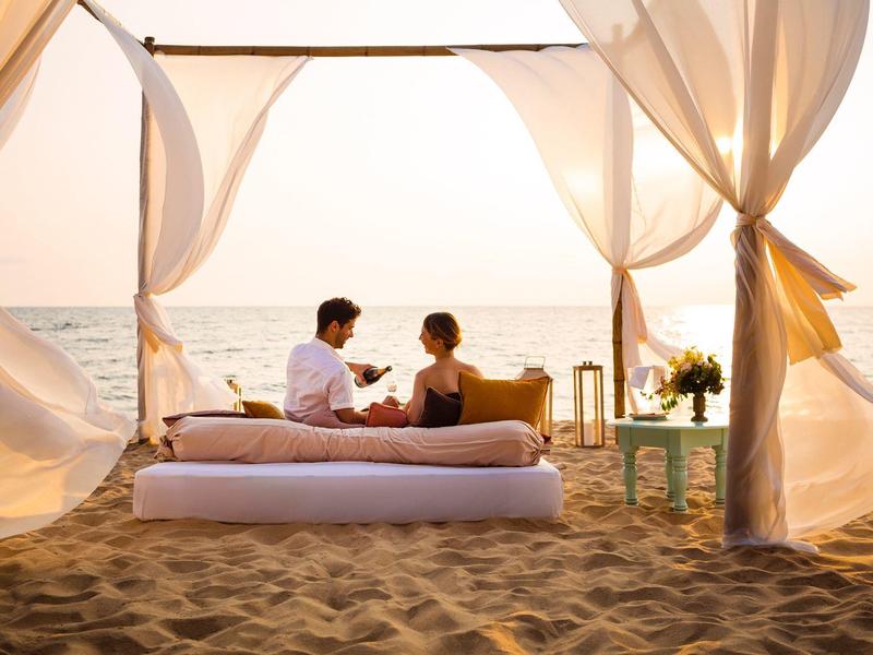 Couple enjoying sunset on the beach under an open canopy with white curtains.