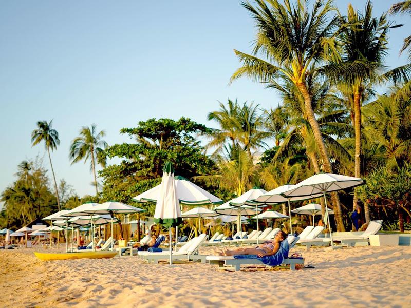 Beach with lounge chairs and white umbrellas surrounded by palm trees in sunlight.
