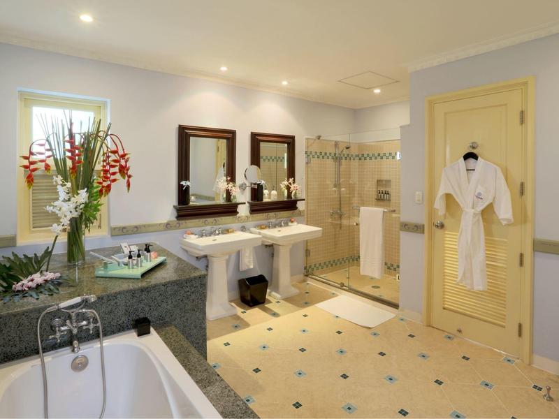 Bright bathroom with double sinks, large bathtub, flowers, and bathrobes hanging on the door.