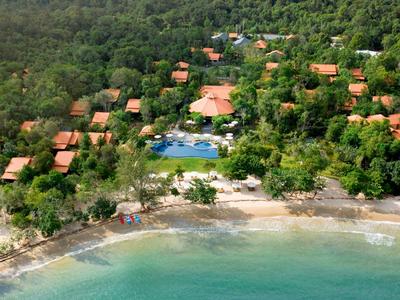 Aerial view of beachfront villas surrounded by lush forest and a central pool near the shoreline.
