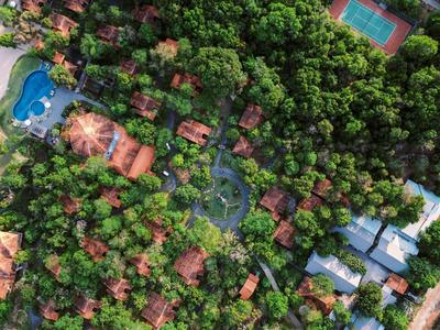 Aerial view of a lush green resort with wooden cabins, swimming pools, and a tennis court.