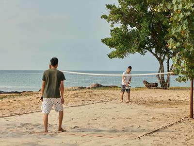 Zwei Männer spielen Beachvolleyball am Strand mit Blick aufs Meer und Bäume im Hintergrund.