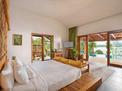 Bright hotel room with large bed, wooden accents, seating area, and doors opening to a terrace with pool view.