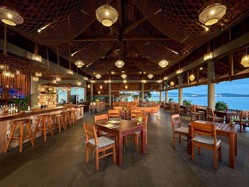 Cozy hotel restaurant with wooden tables, chairs, and warm hanging lights by the sea at sunset.