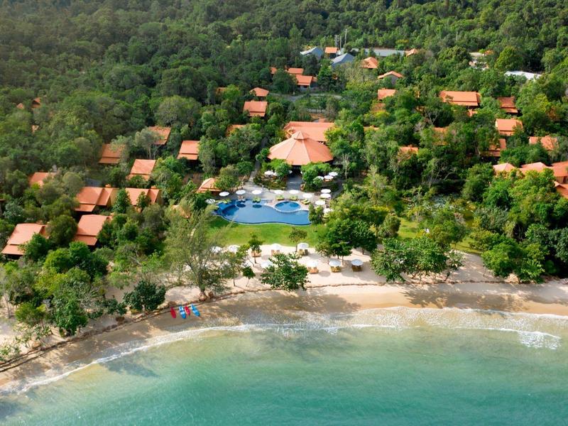 Aerial view of beachfront villas surrounded by lush forest and a central pool near the shoreline.