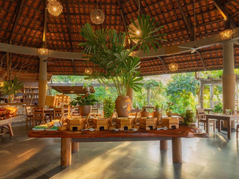 Open-air restaurant with wooden furniture, tropical plants, and warm lighting under a thatched roof.