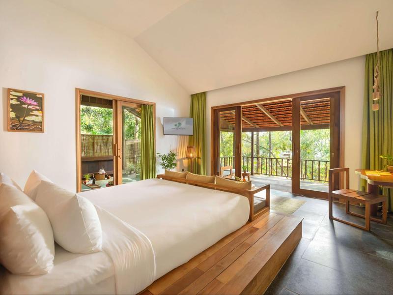 Spacious hotel room with large bed, wooden furniture, and balcony overlooking greenery.