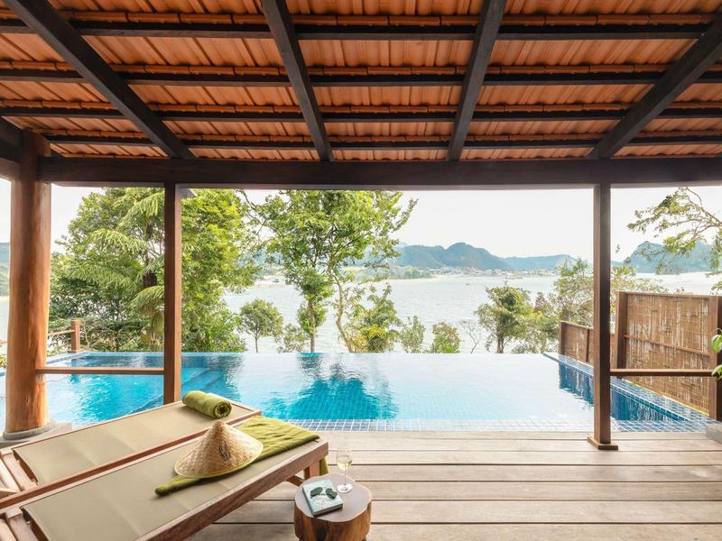 Relaxing infinity pool view from a wooden deck with lounge chairs under a tiled roof.