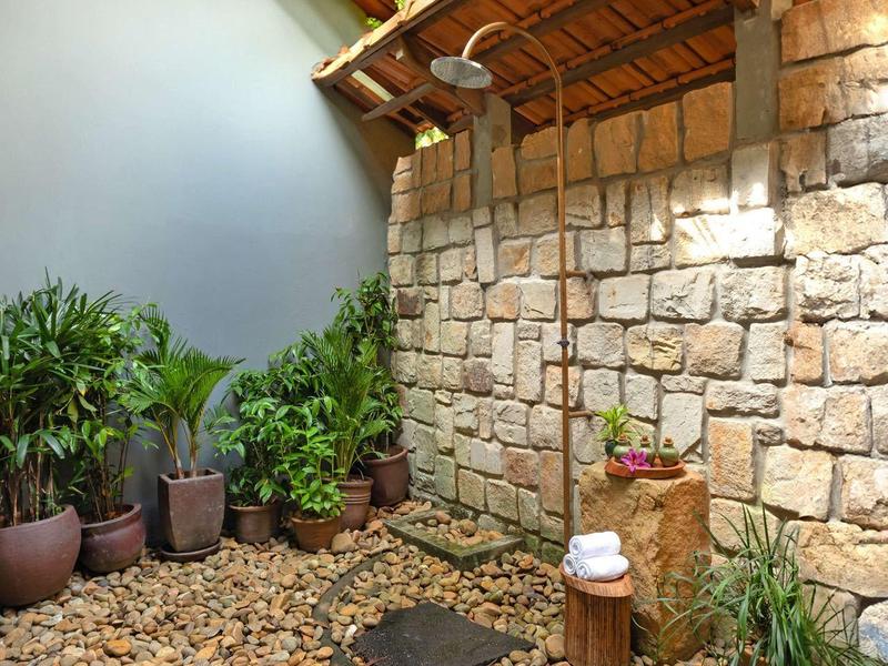 Cozy outdoor shower area with stone wall, wooden roof, and pebble ground with stepping stones.