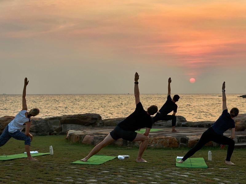Four people practicing yoga on mats by the sea during a colorful sunset.