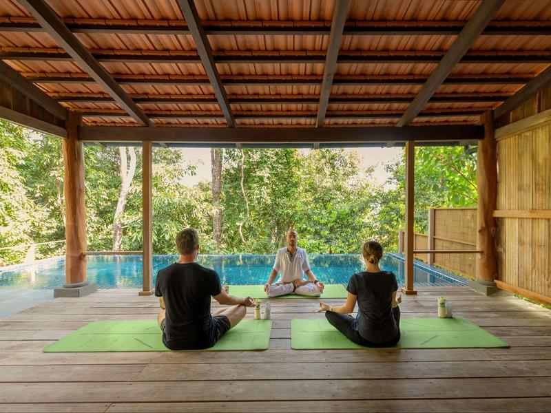 Three people seated on yoga mats in an open wooden pavilion with a forest and pool view.