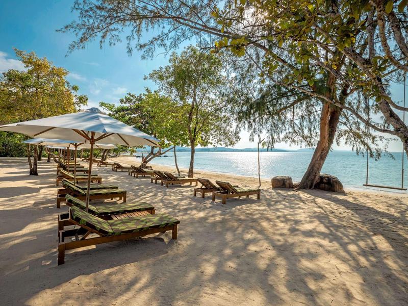 Sandy beach with lounge chairs and umbrellas shaded by trees near calm blue water.