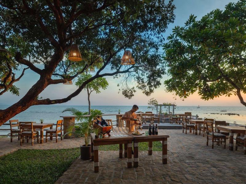 Outdoor dining area under trees with wooden tables and sea view at sunset.