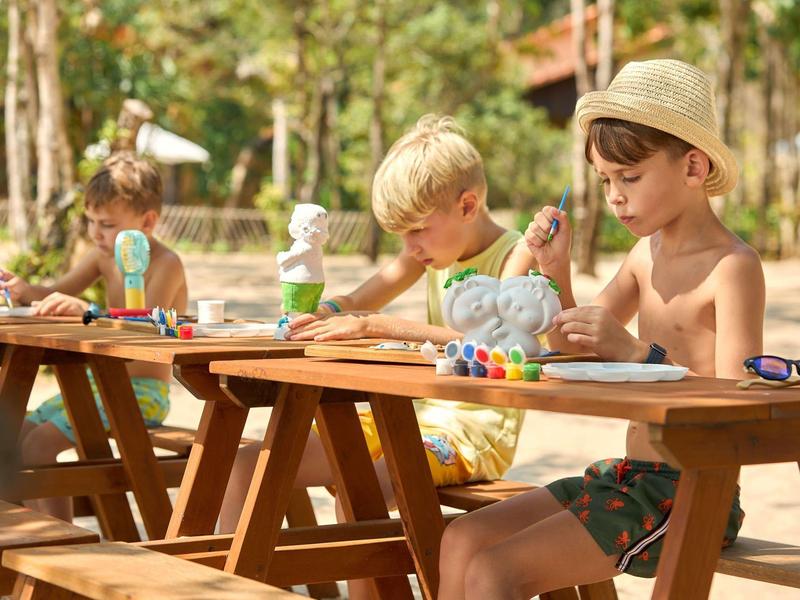 Children sitting at picnic tables outdoors, painting Easter eggs on a sunny day.