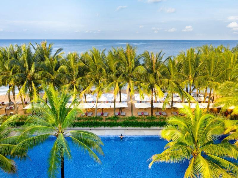 Piscina azul con palmeras y vista a una playa de arena y el mar bajo un cielo despejado.