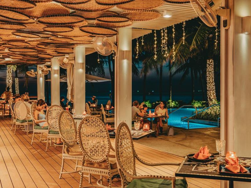 Acogedora zona al aire libre con alfombras, muebles de rattán y chimenea junto a piscina y palmeras de noche.