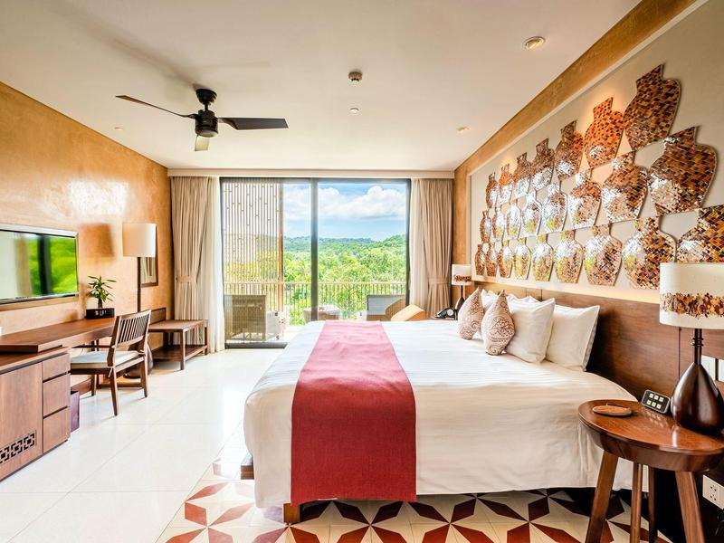 Habitación de hotel luminosa con cama grande, puerta terraza y vista a la montaña, tonos cálidos.
