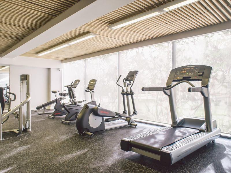 Gimnasio luminoso con cintas de correr, elípticas y bicicletas estáticas frente a grandes ventanas.