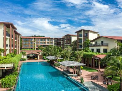 Großes Resort mit Pool, umgeben von Gebäuden und tropischem Grün unter blauem Himmel.
