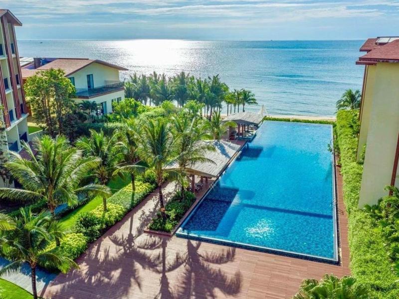 Blick auf blauen Pool, umgeben von Palmen und Gebäuden, mit Meer im Hintergrund bei Tageslicht.