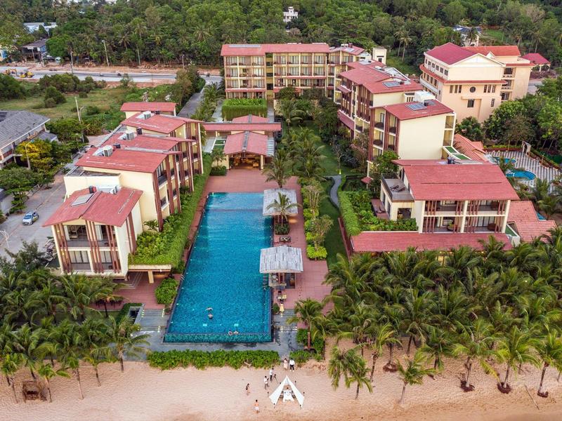 Helles Resort mit Pool, umgeben von grünen Palmen direkt am Sandstrand. Mehrere Gebäude mit roten Dächern.