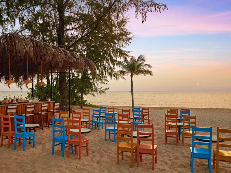 Bunte Holzstühle auf Sand am Strand mit Palmen und ruhigem Meer im Sonnenuntergang.