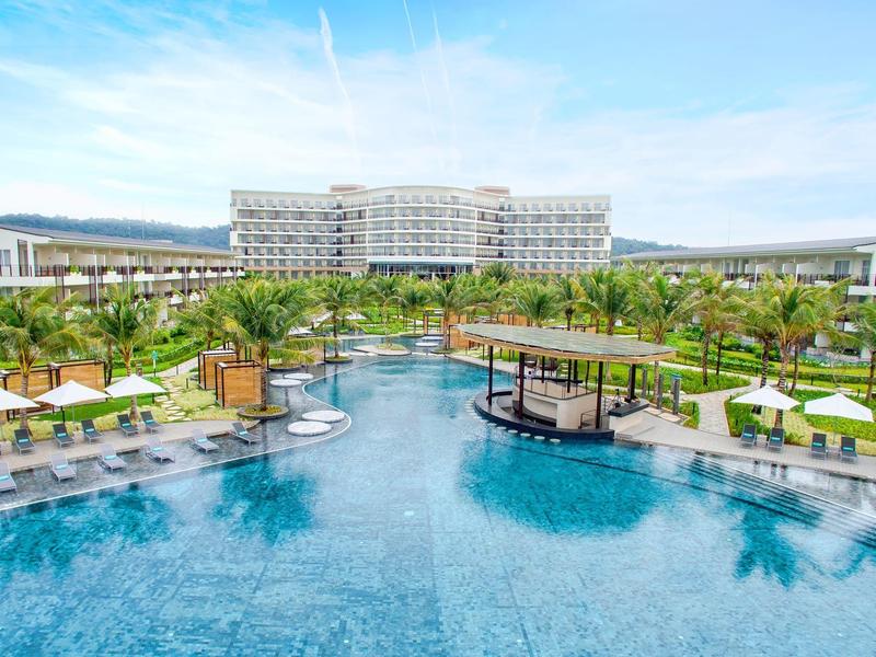 Großer blauer Pool mit Palmen und Sonnenschirmen vor einem modernen Hotelgebäude unter blauem Himmel.