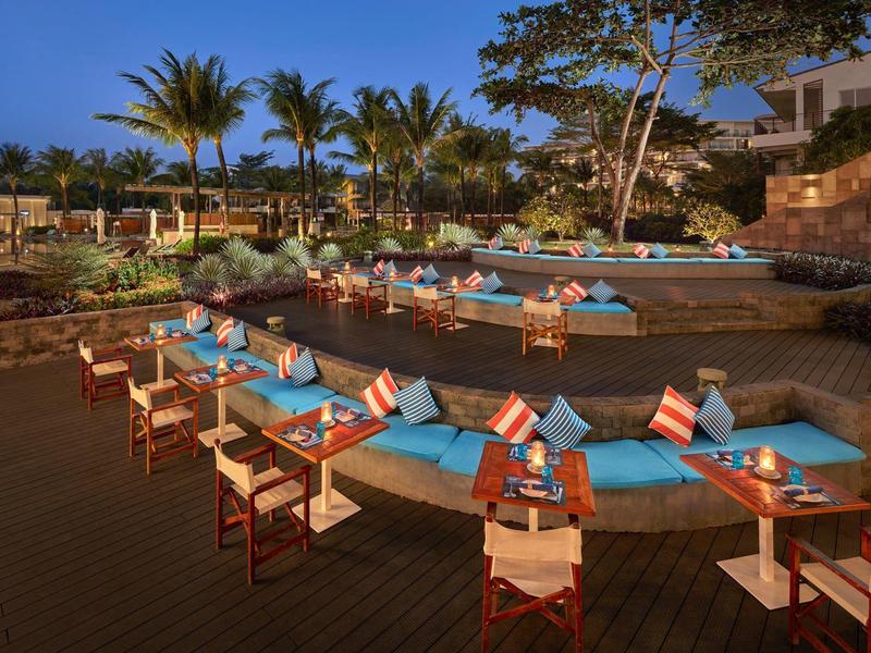 Gemütliche Terrasse mit blauen Sitzpolstern, Holztischen und weißen Stühlen bei Abenddämmerung.