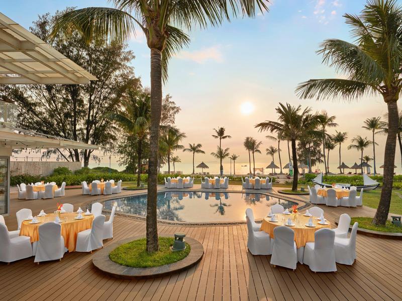 Luxuriöse Terrasse mit runden Tischen, dekorierten Stühlen, Pool und Palmen im Sonnenuntergang.