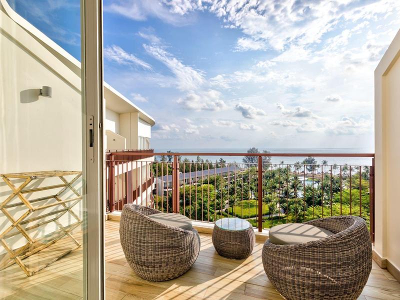 Balkon mit Rattanmöbeln, kleinem Tisch, Holzgeländer und Blick auf Häuser und Meer bei Sonnenschein.