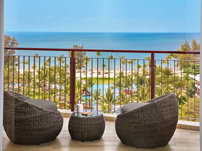 Balkon mit zwei Rattansesseln, kleinem Tisch, Blick auf Palmen, Pool und das blaue Meer im Hintergrund.