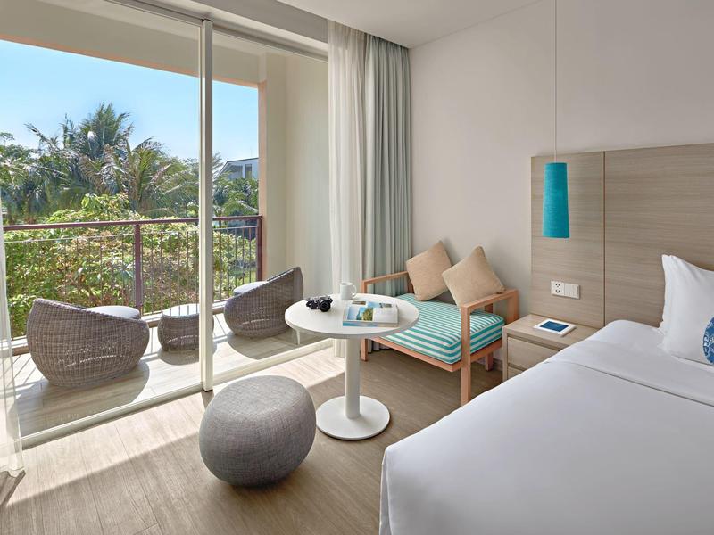 Helles Hotelzimmer mit Doppelbett, Sitzbereich, Glas-Schiebetür und Balkon mit Stühlen.