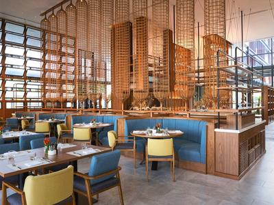 Modern eingerichtetes Restaurant mit blauen und gelben Stühlen, Holztischen und großen Fenstern.