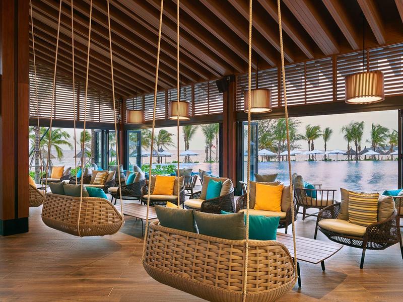 Gemütlicher Lounge-Bereich mit hängenden Korbsesseln, bunten Kissen und Sicht auf Palmen und Wasser.