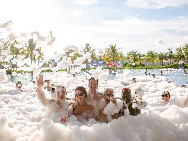 Gruppe von Menschen in einem schaumgefüllten Pool bei sonnigem Wetter, umgeben von Palmen.