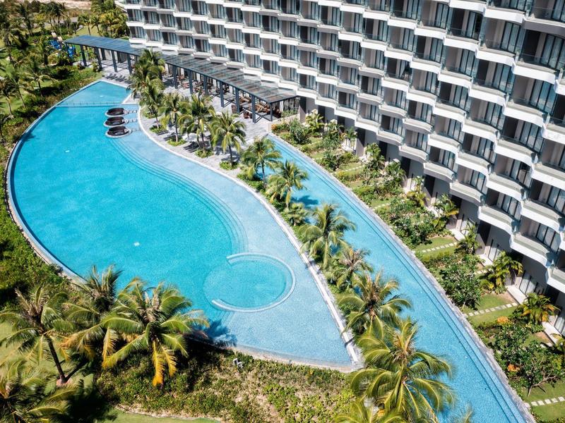 Großer, geschwungener Pool neben einem Hotel mit Balkonen und Palmen.