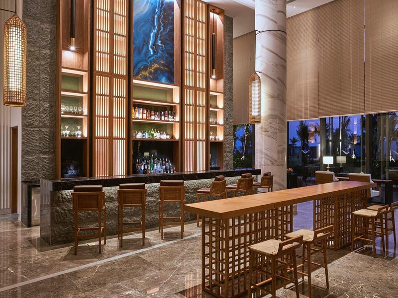 Moderner Hotelbarbereich mit hohem Deckengewölbe, beleuchtetem Tresen und Holzstühlen in hellem Ambiente.