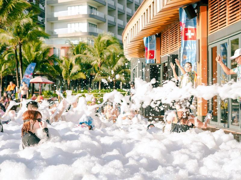 Menschen genießen eine große Schaumparty im Freien bei Sonnenschein vor einem Gebäude.