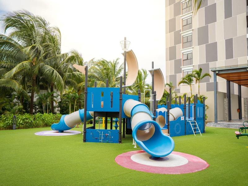 Bunter Spielplatz mit Rutschen auf grünem Rasen vor Palmen und modernem Gebäude.