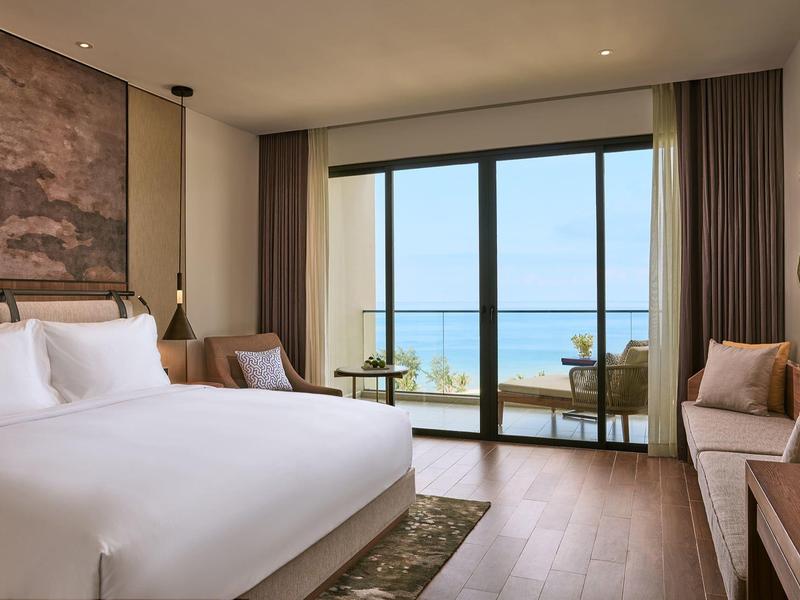 Helles Schlafzimmer mit großem Bett, Holzboden, Balkon und Meerblick durch bodentiefe Fenster.