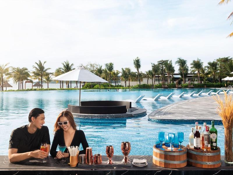 Zwei Personen unterhalten sich an einer Bar am Pool mit Palmen im Hintergrund.