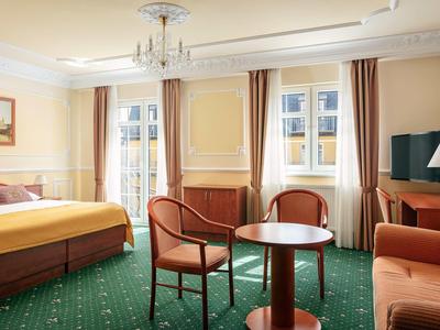 Bright hotel room with double bed, two chairs, table, and sofa on green carpet.